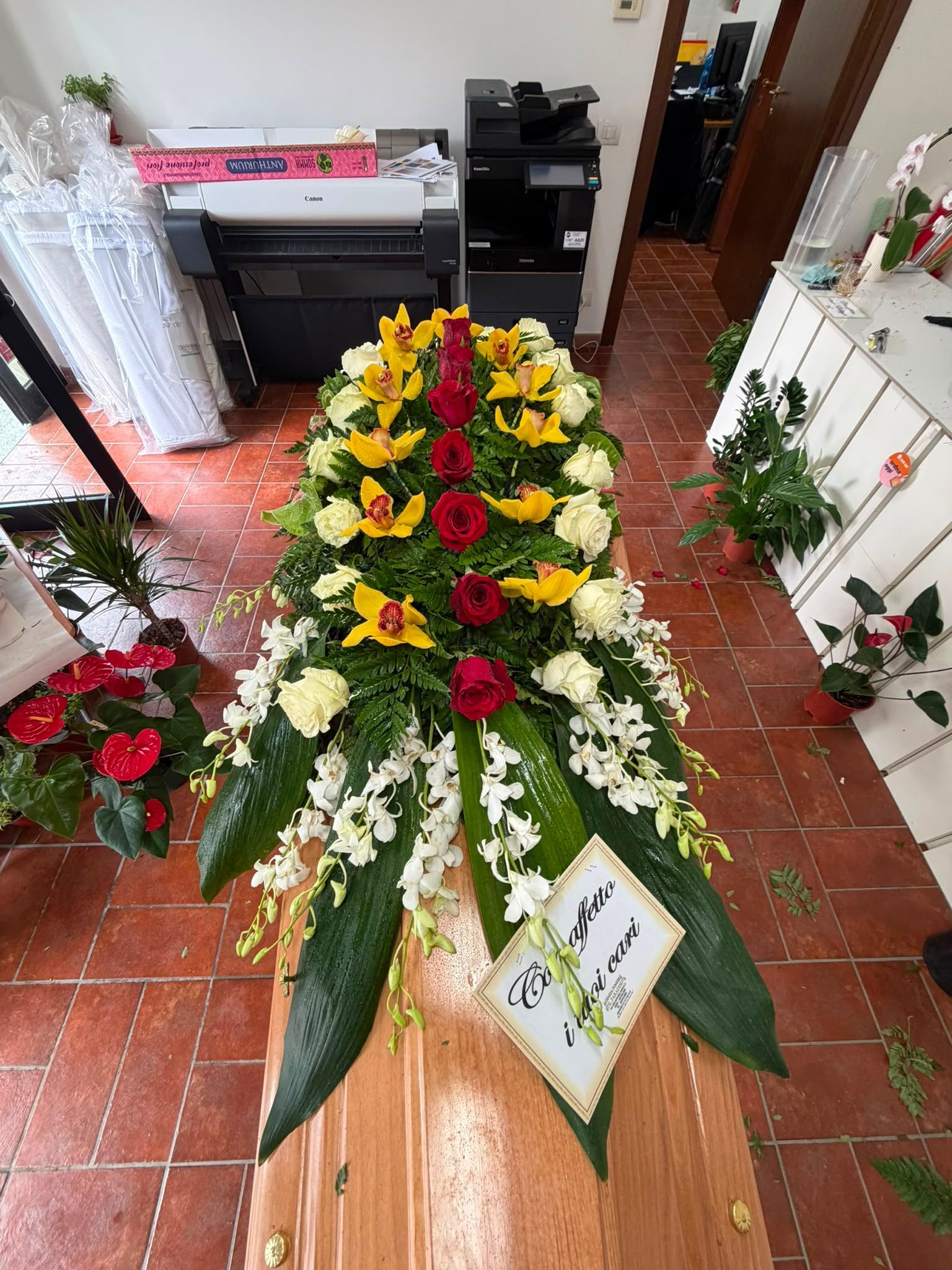 Corona funebre con fiori rossi, gialli e bianchi, su un supporto al chiuso, sopra un pavimento piastrellato