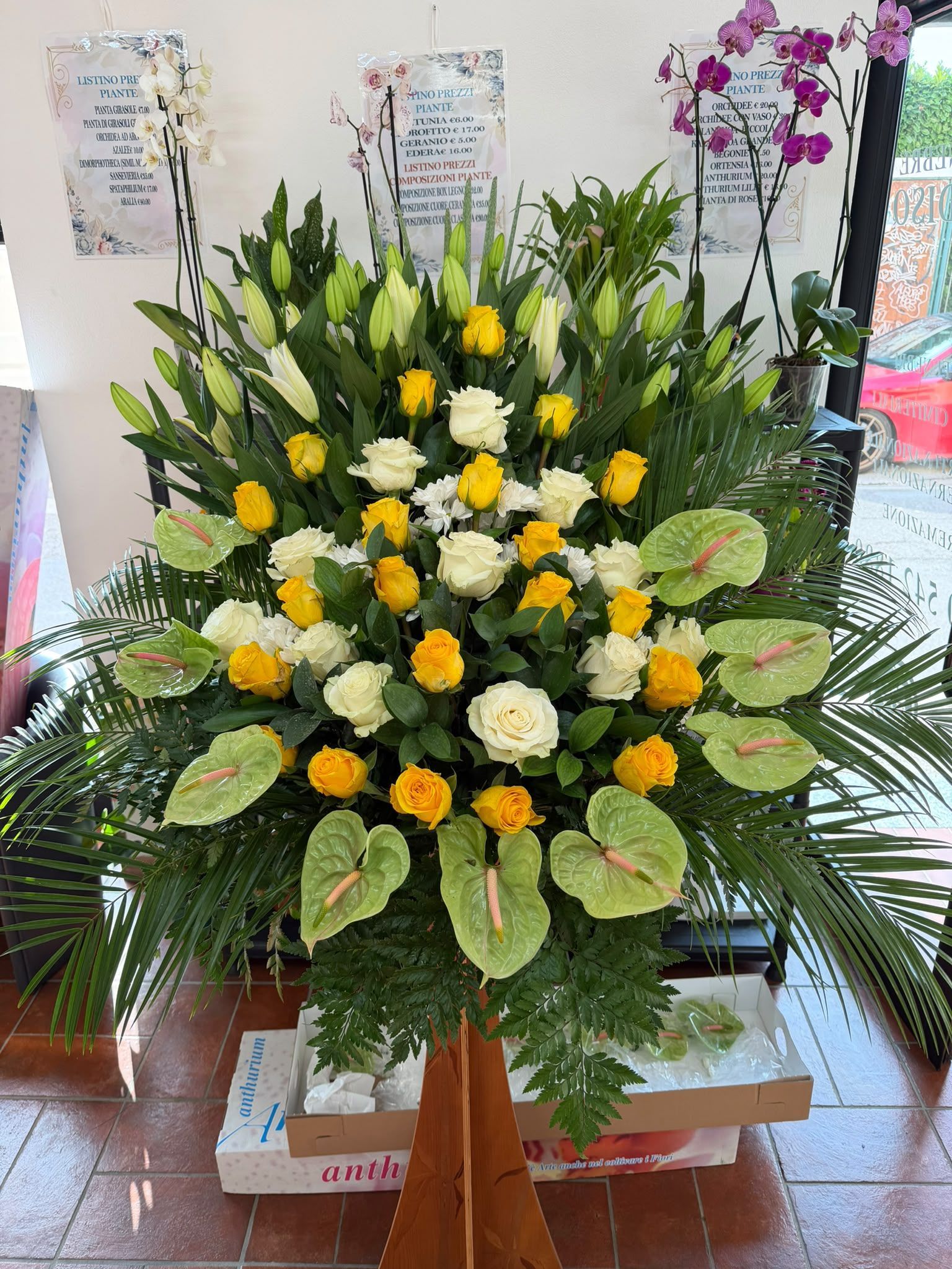 Grande composizione floreale funebre con fiori bianchi e gialli, foglie verdi e orchidee rosa, in un ambiente interno