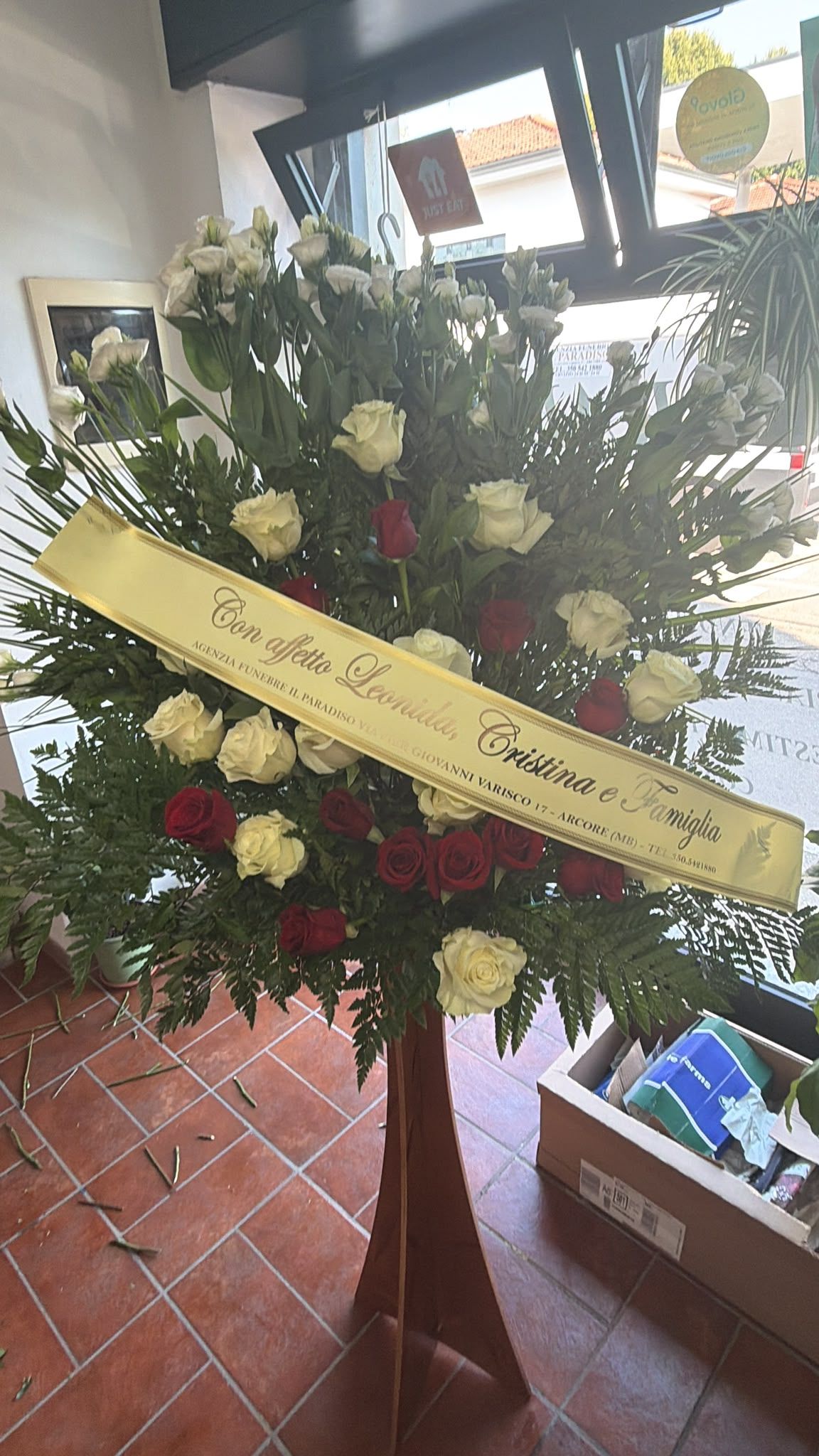 Corona funebre floreale con fiori bianchi e rossi e un nastro beige su un supporto, in un ambiente interno