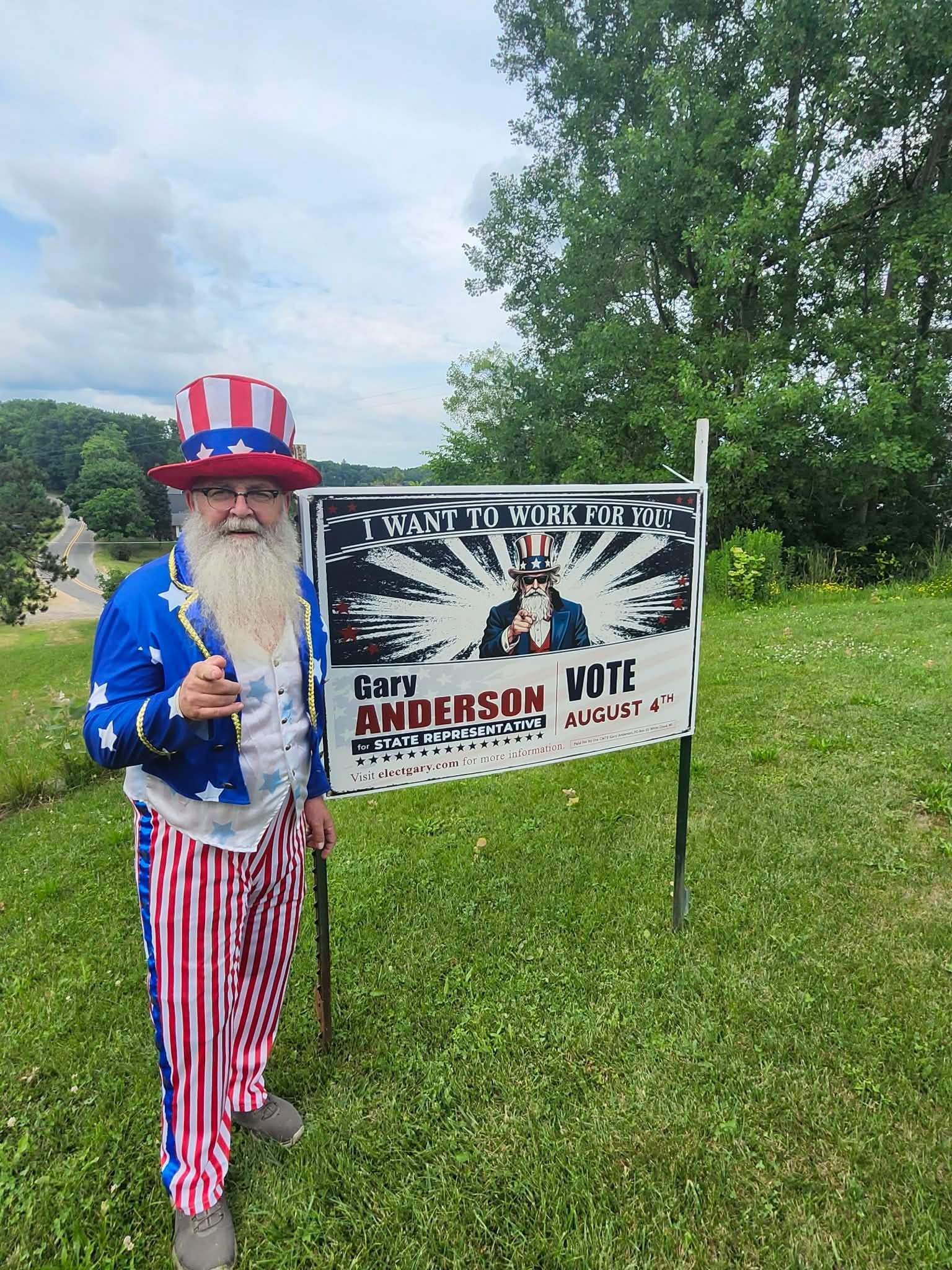 Gary in red-white-blue outfit beside a protest sign on a grassy roadside
