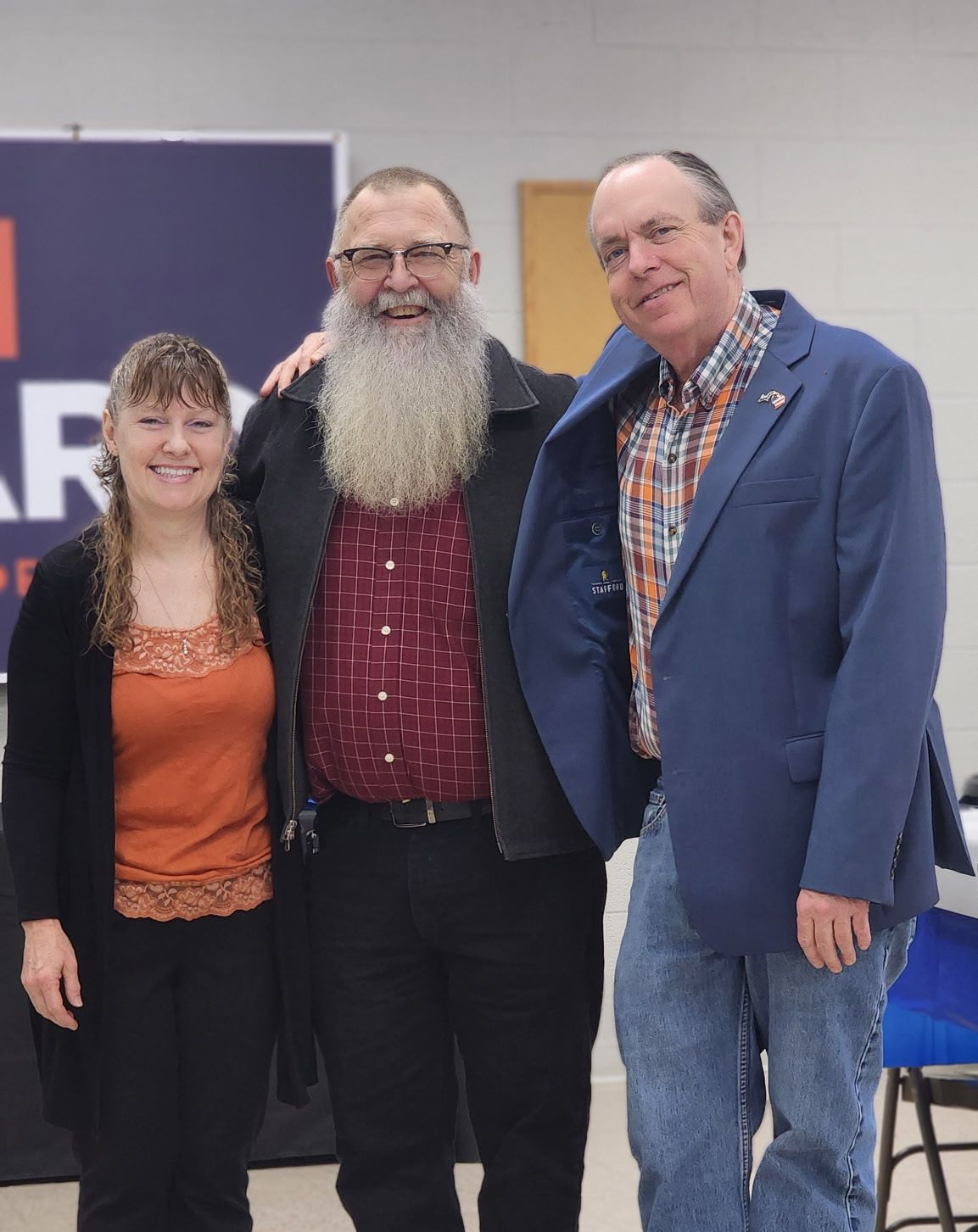 Three people smiling together: woman, man with long beard, man in blue jacket.