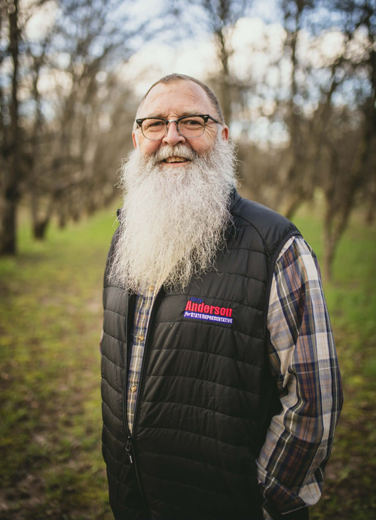 Man with long white beard stands in snow, hands in pockets. Trees in background.
