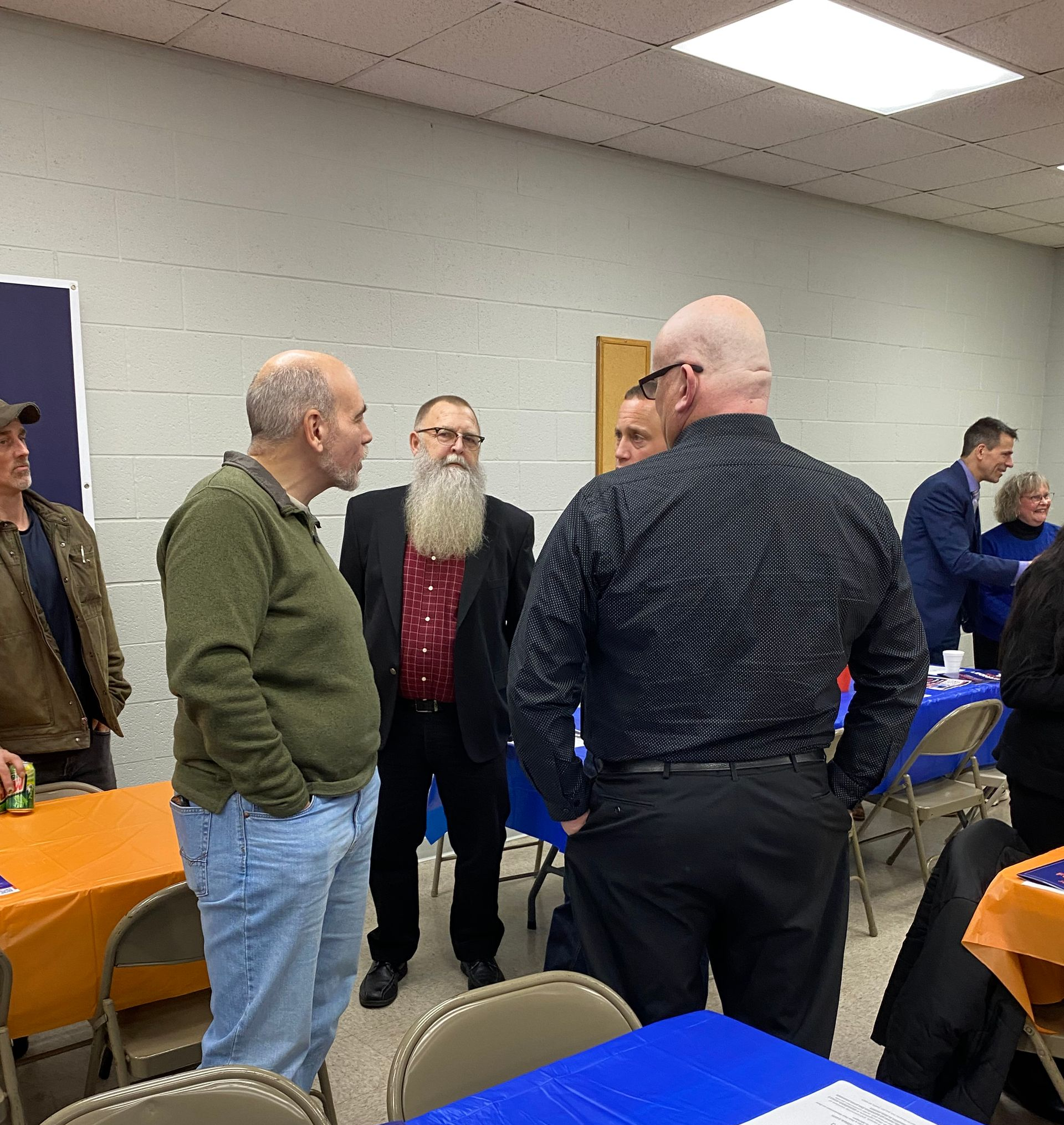 People talking at an event, orange and blue tables, a man with a long beard.