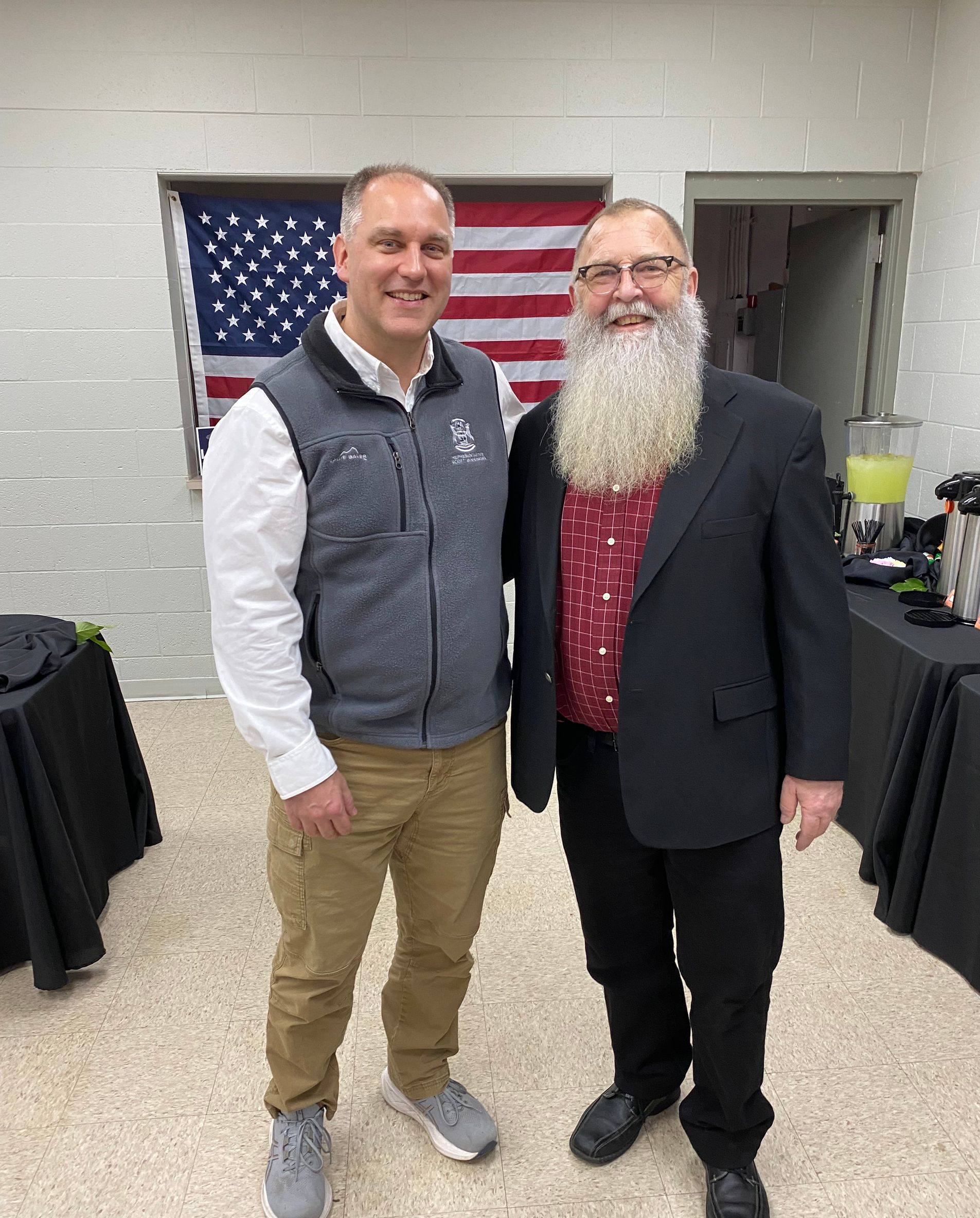 Two men pose in front of an American flag; one in a vest, other with a long white beard.