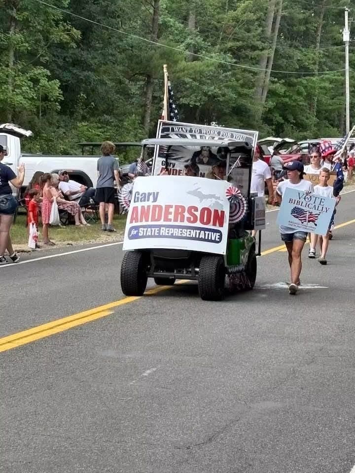 Parade float with “ANDERSON” campaign sign on a street, with people walking alongside and trees in the background