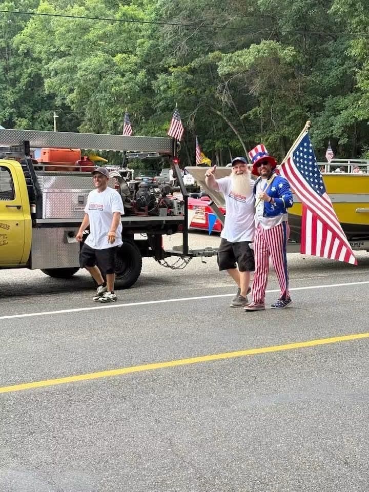 Parade participants walking beside a truck, one holding a large American flag on a tree-lined road.
