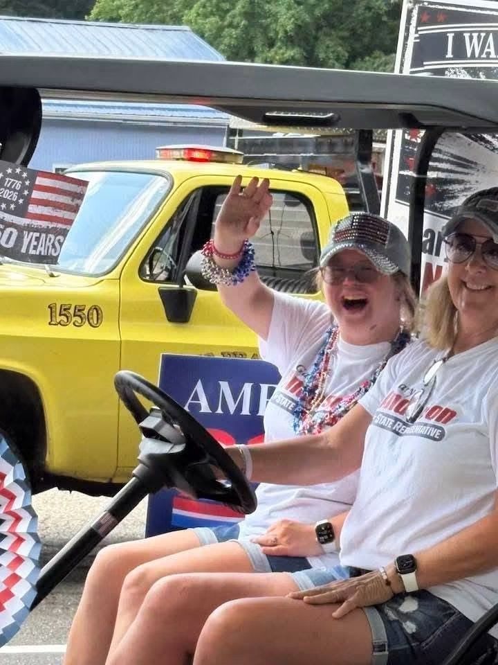 Two smiling people wave from a yellow AMR golf cart at an outdoor event.