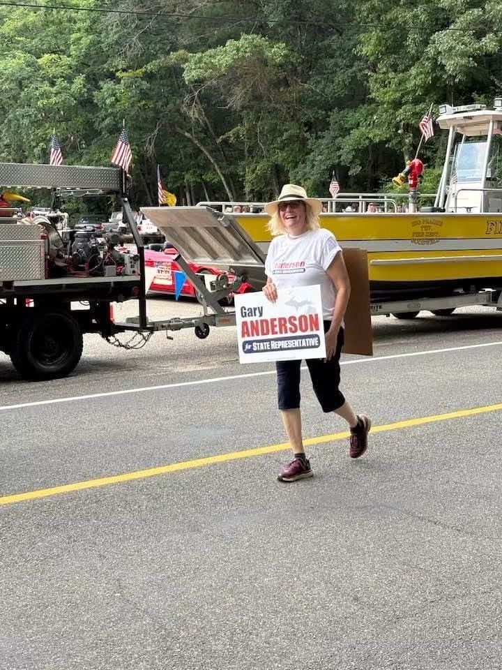 Person walking on roadside holding a campaign sign beside a truck and yellow boat