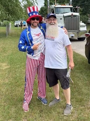 Two men posing outdoors, one in a red-white-blue patriotic costume and the other in a white T-shirt, near a truck.