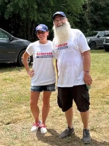 Two people in matching white shirts and blue caps standing outdoors beside parked cars and grass.