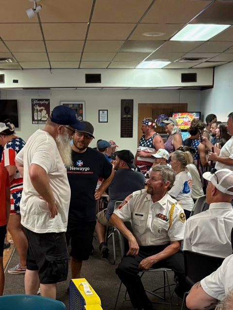 Community meeting in a crowded room, with a man speaking to a seated audience near a yellow object in the foreground.