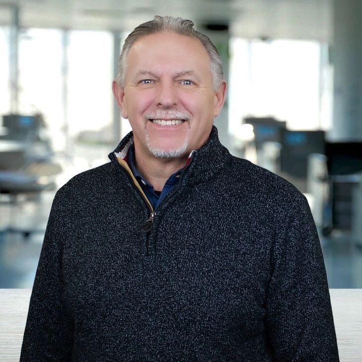 Man with graying hair and beard smiles, wearing a navy blue sweater; blurred background.