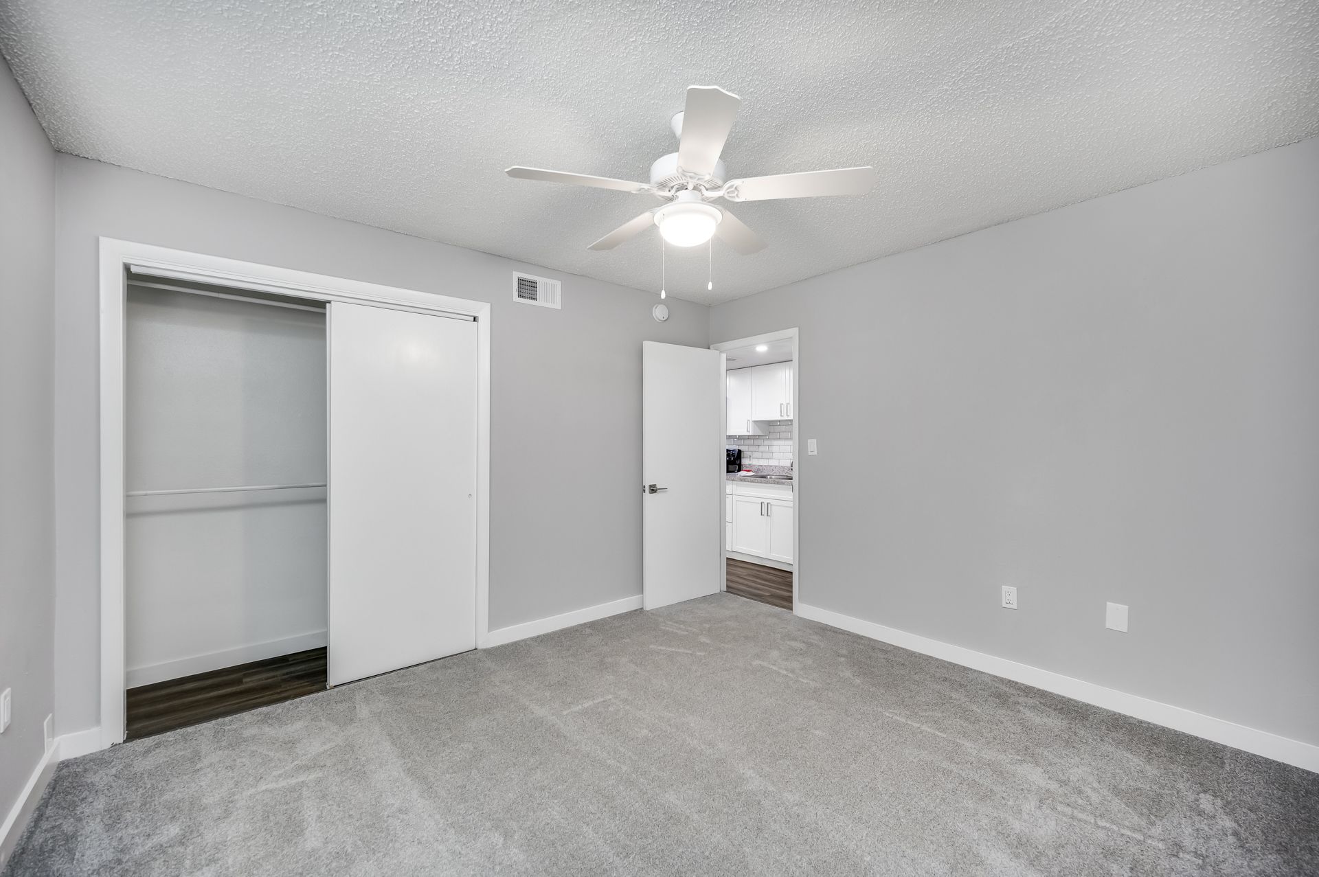 Empty bedroom with gray carpet, walls, and closet. Door leads to kitchen, ceiling fan present.