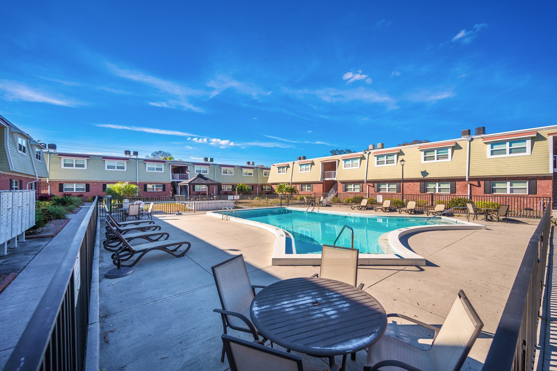 Outdoor pool surrounded by townhouses, lounge chairs, and a table on a sunny day.