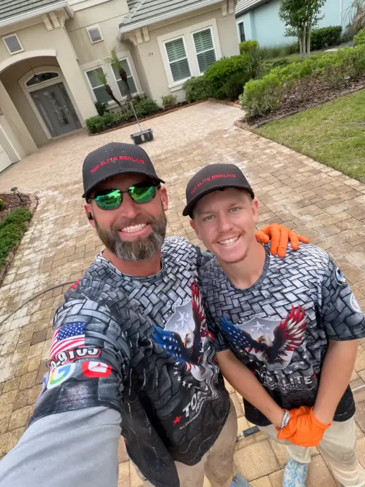 Two men in matching shirts and caps smiling on a brick driveway in front of a house