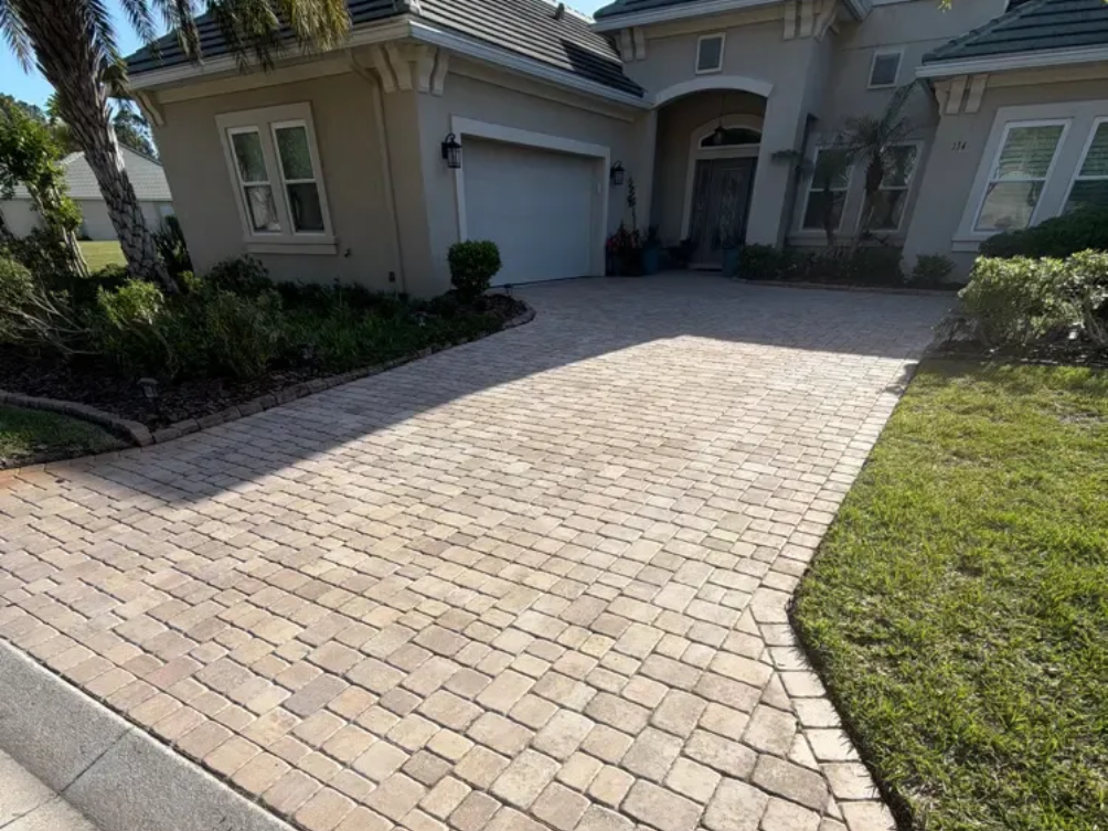 Brick-paved driveway leading to a beige house with a garage and front entryway.