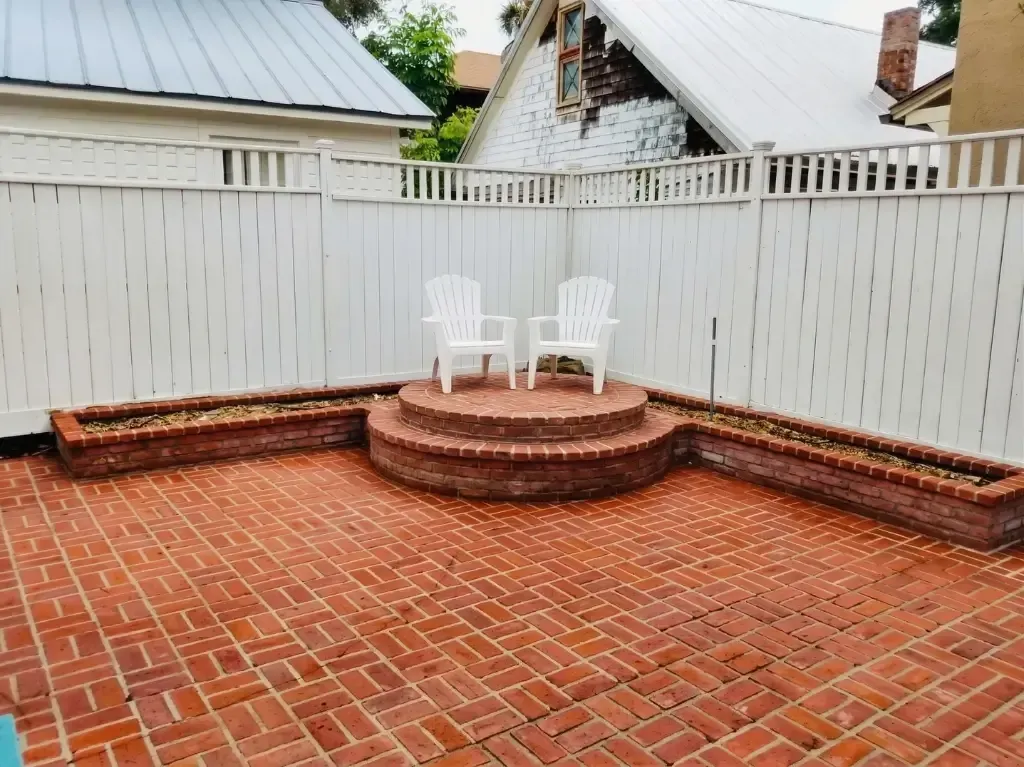 Brick patio with curved steps, white fence, and two white chairs in a small backyard.