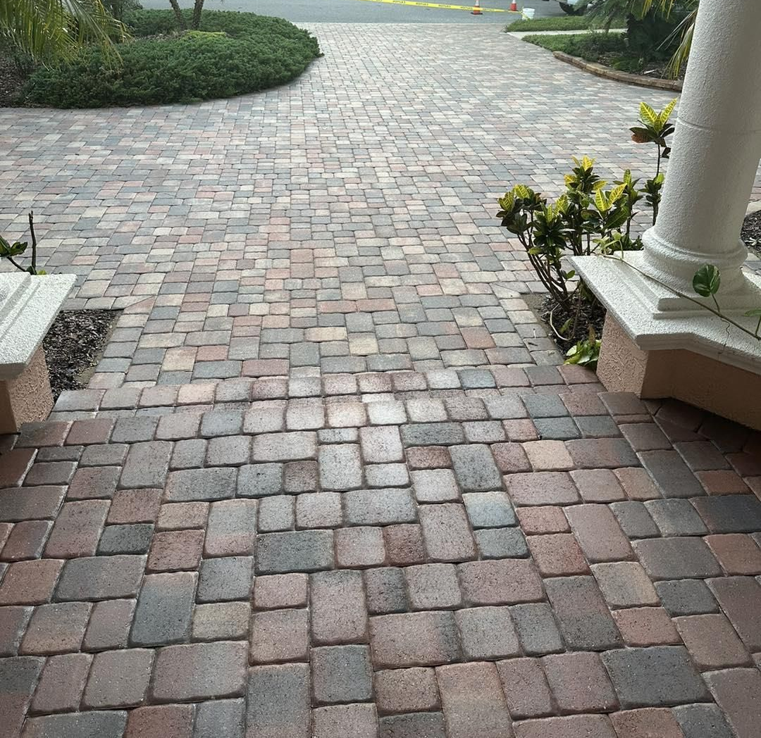 Wet brick walkway leading through a garden toward a road, with benches and white columns on the right