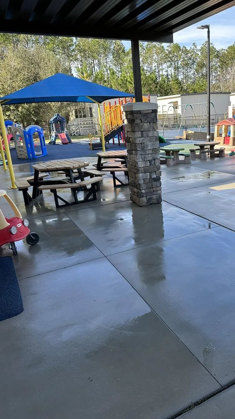 #1 Master Certified concrete cleaning and sealing in Saint Augustine! A clean concrete commercial patio area featuring picnic tables under a canopy, with a playground visible in the background.
