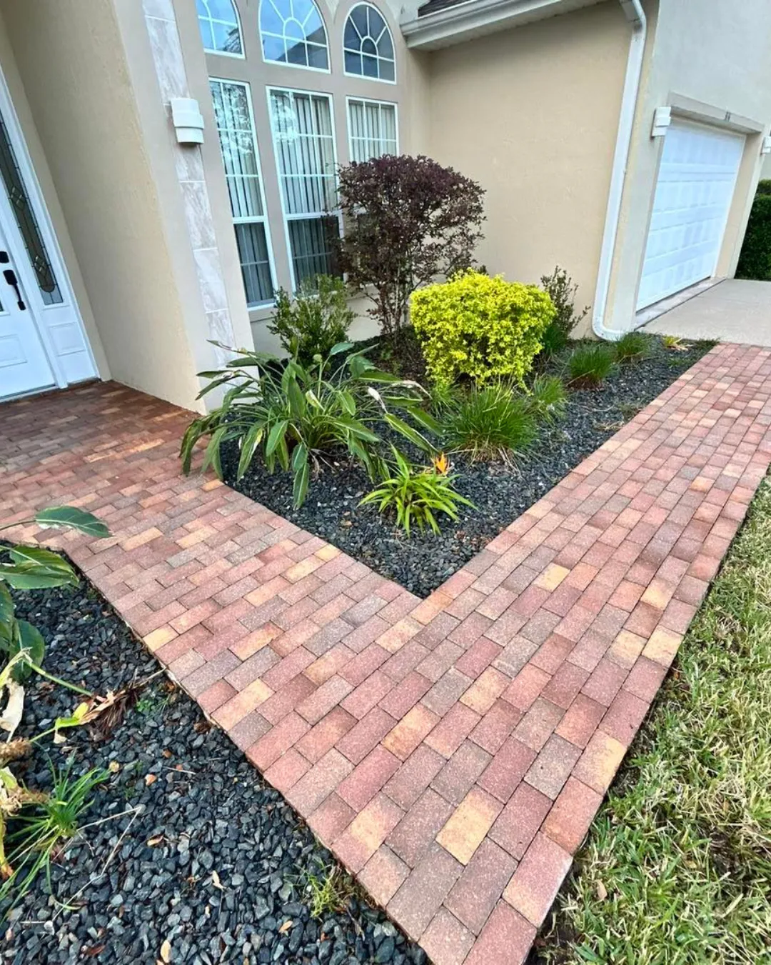 Brick walkway edging a front yard garden with green and yellow shrubs beside a house entrance