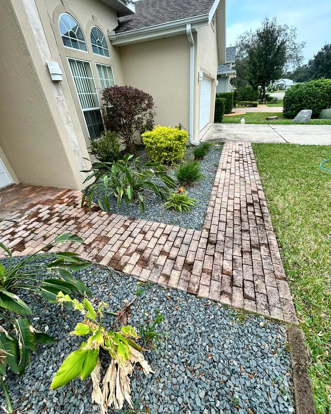 Brick and gravel front walkway beside a beige house with landscaped plants and shrubs
