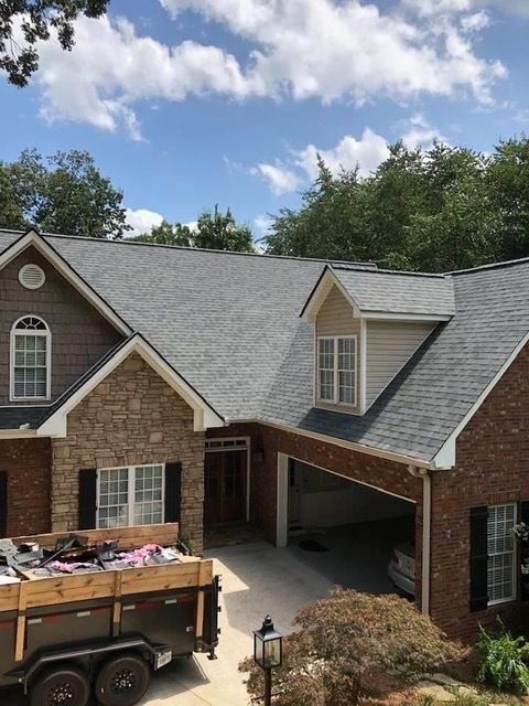 House with new gray roof, brick and stone exterior, and a trailer parked in the driveway under a sunny sky.