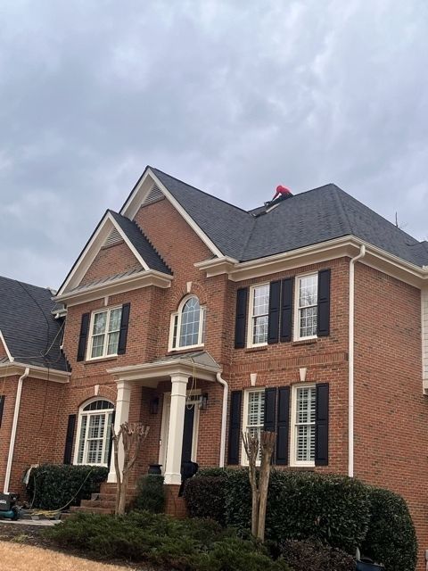 Brick house with a dark roof and a person on top wearing a red jacket, cloudy sky.