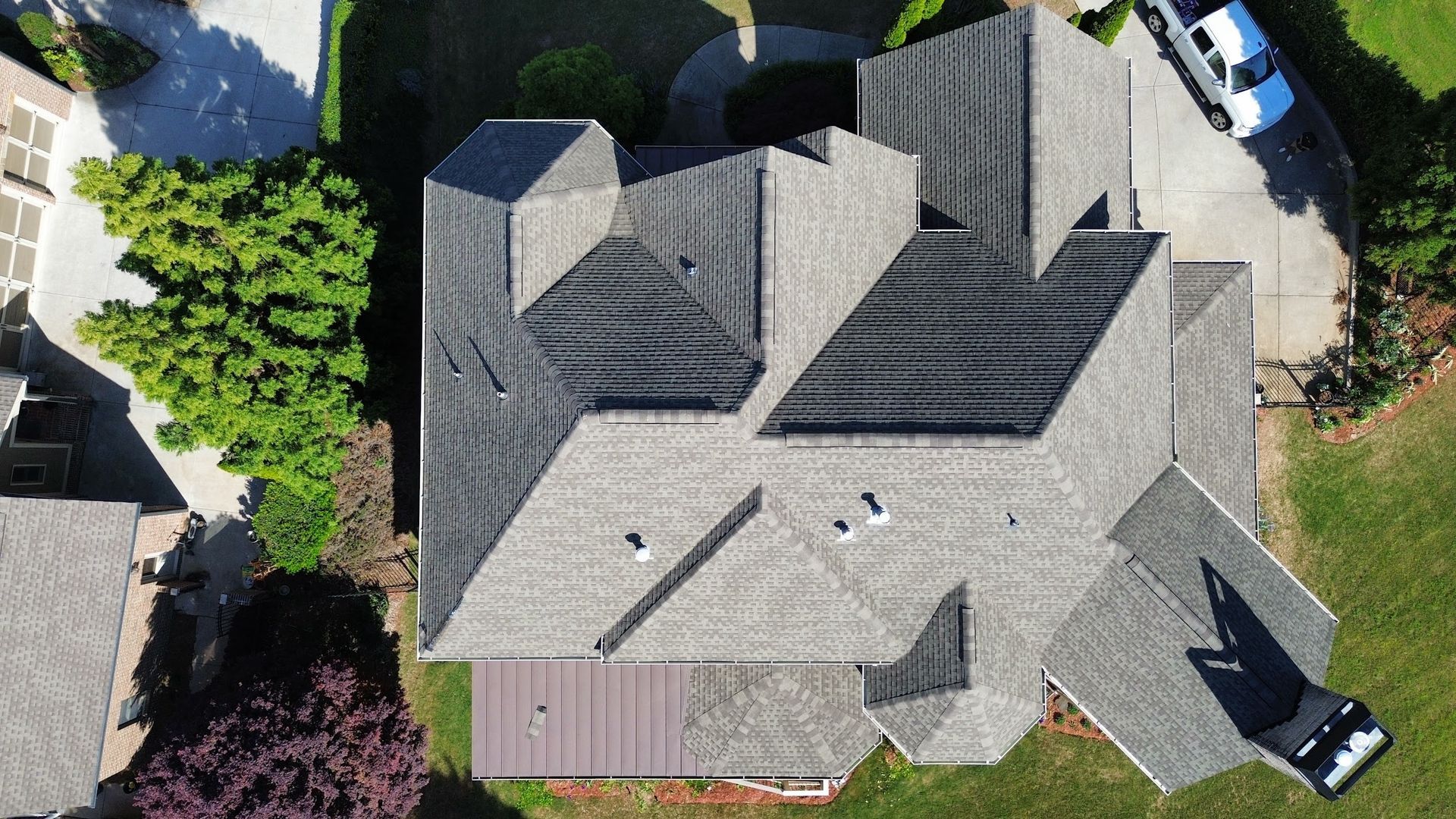 Aerial view of a gray shingle roof with multiple gables on a house surrounded by green trees and grass.
