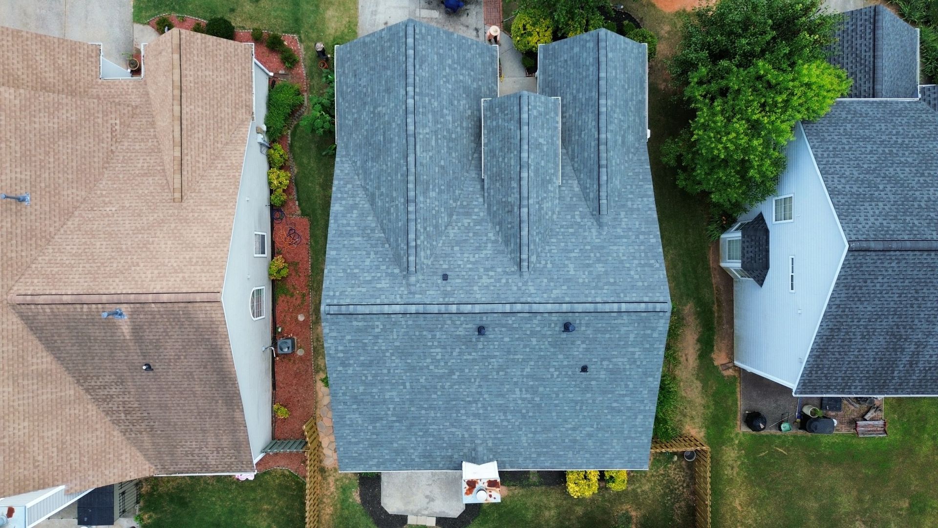 Overhead view of a house with a gray shingled roof between two other houses, surrounded by green grass.
