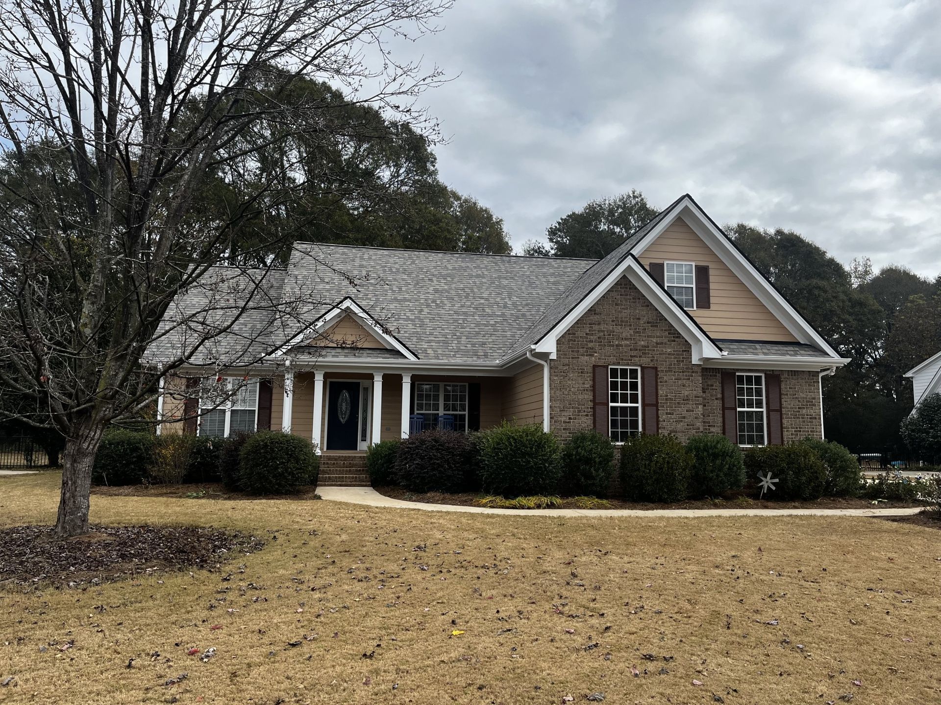 House with brick and tan siding on a cloudy day. Brown bushes line the front. Bare tree in the front yard.