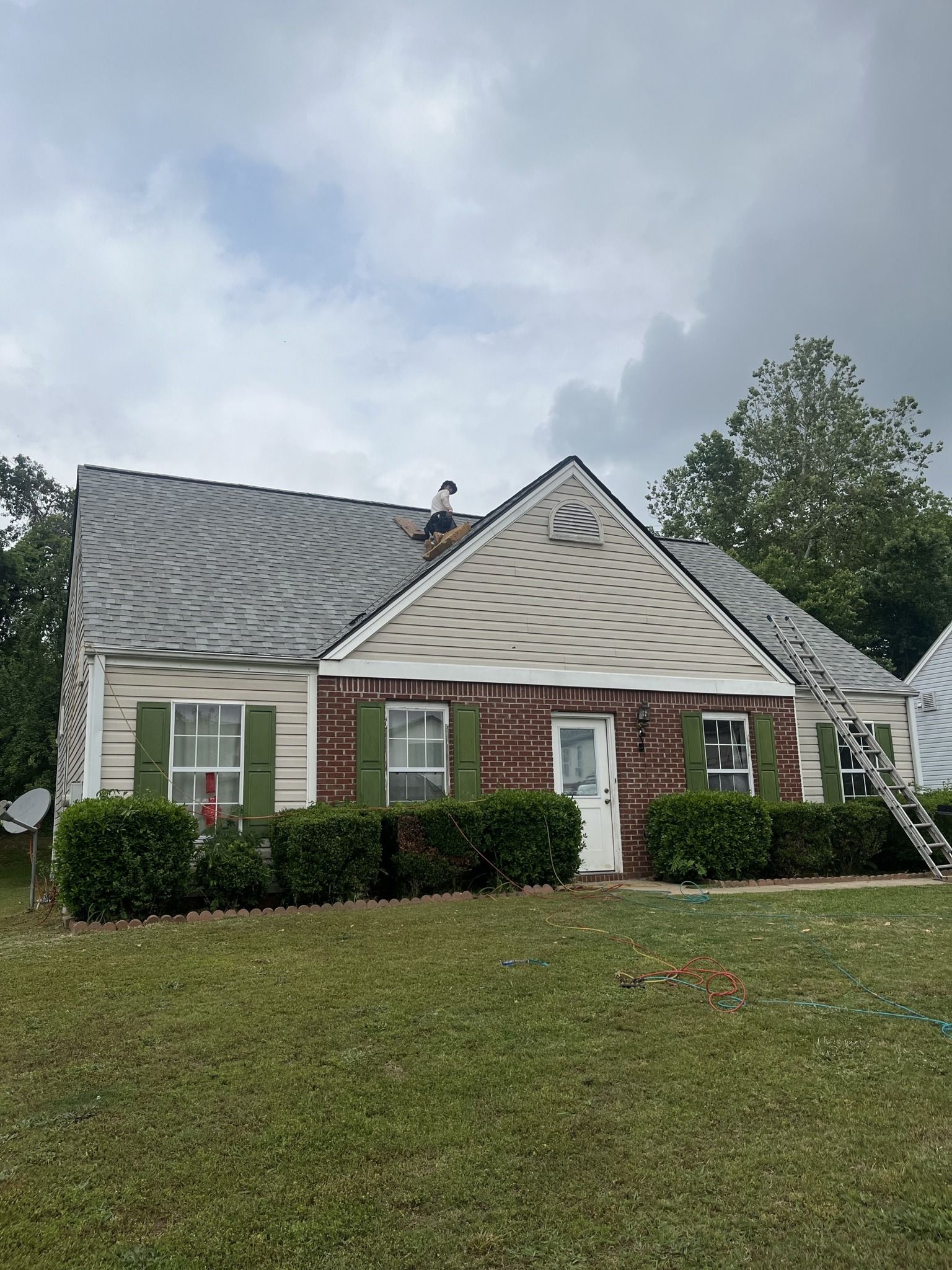 House with missing siding and new roofing; green shutters, white door; grass lawn, cloudy sky.