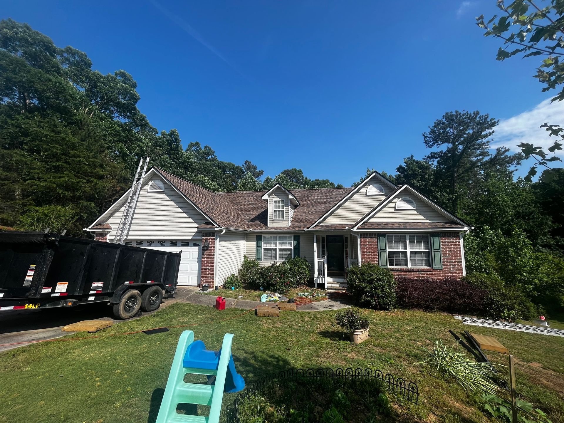 House with damaged roof, surrounded by trees, construction debris, a dumpster, and a blue slide on the lawn.