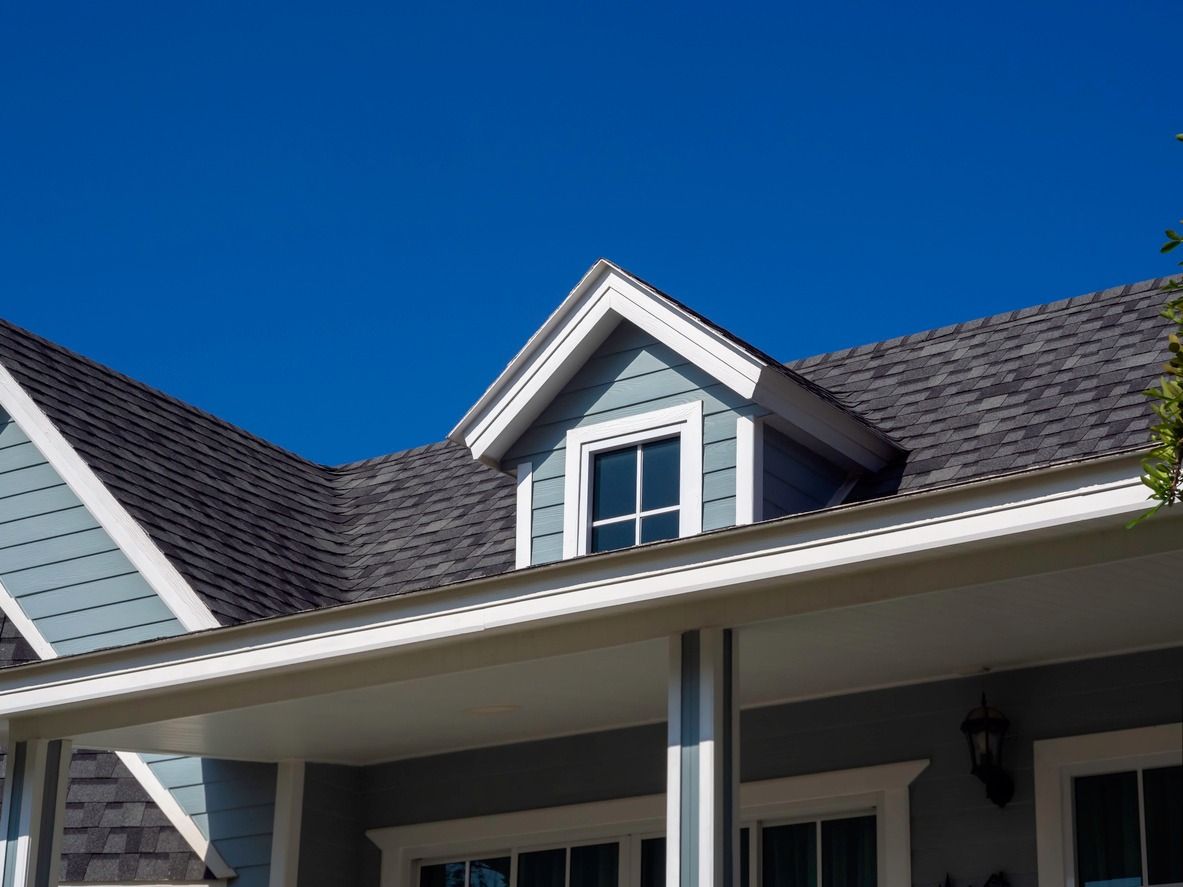 Blue house exterior with dormer window, gray roof, and white trim against a clear blue sky.