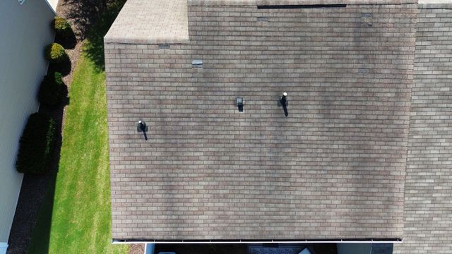 Overhead view of a house roof with weathered shingles, featuring vents and antennas, next to green grass and a white wall.