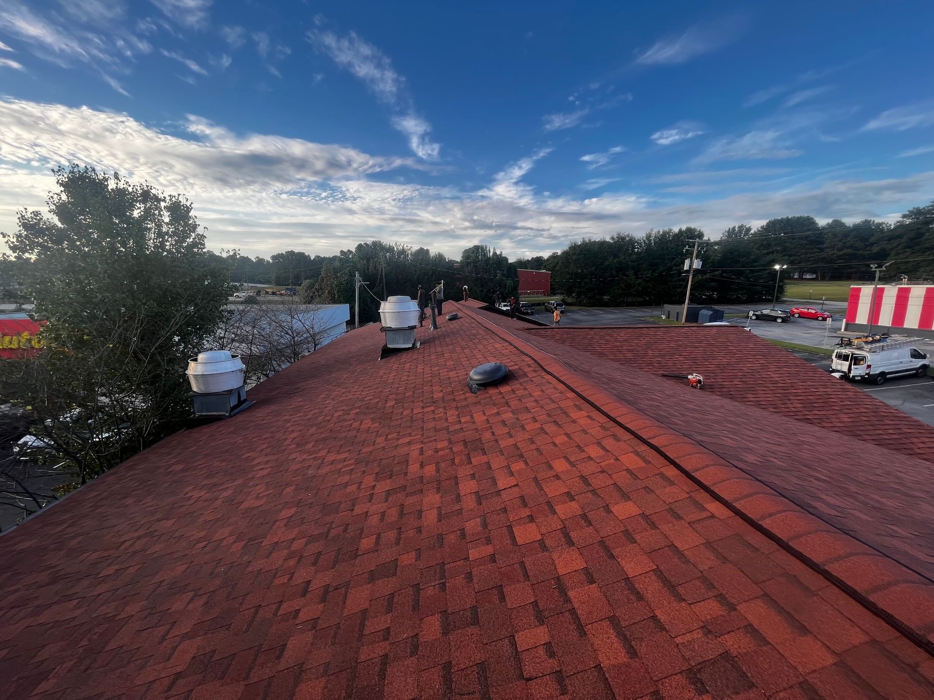 Red shingle roof with vents; trees, buildings, and a partly cloudy blue sky in the background.