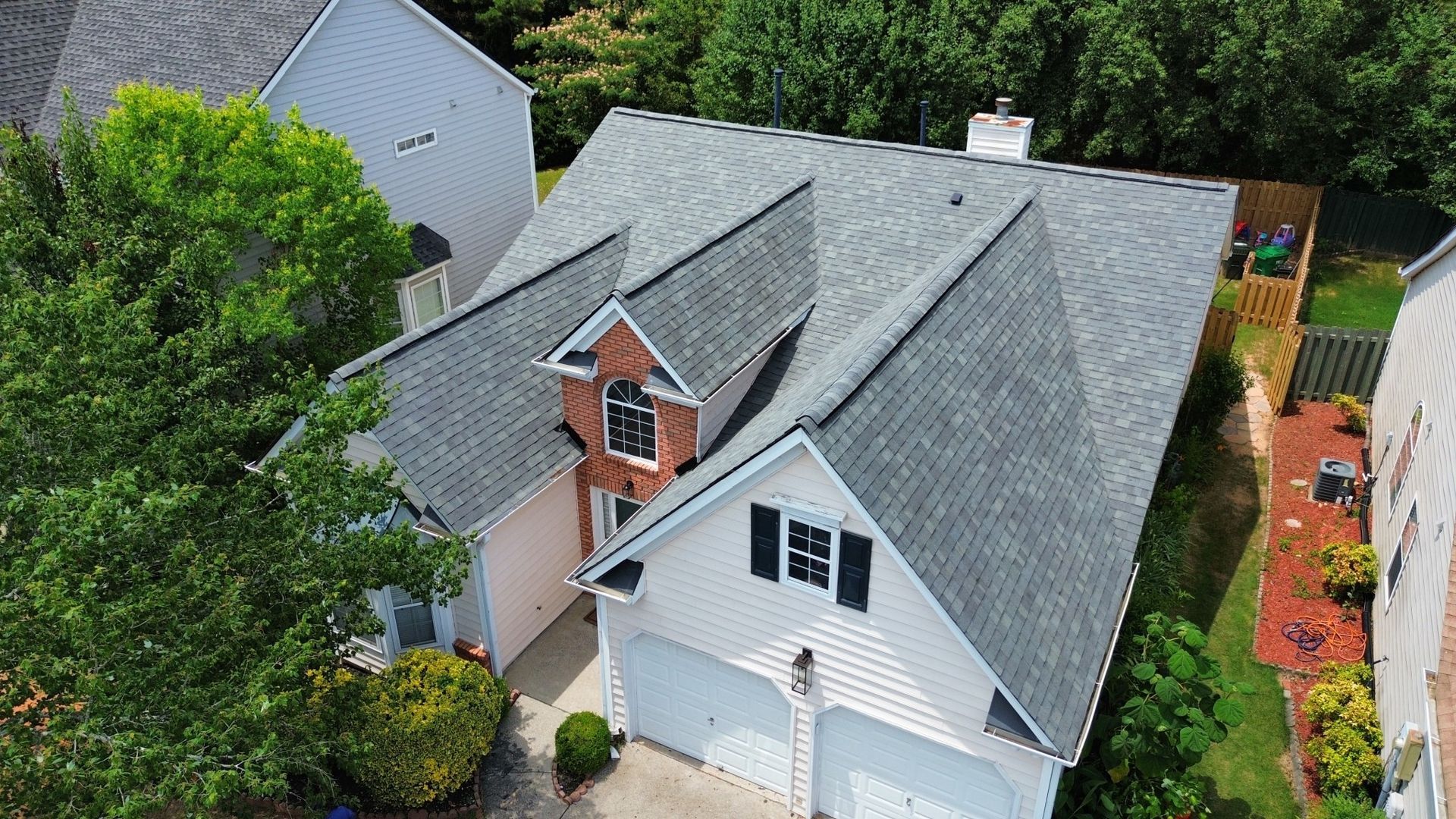 A house with gray roof and white siding, a two-car garage, and surrounded by trees and green grass.