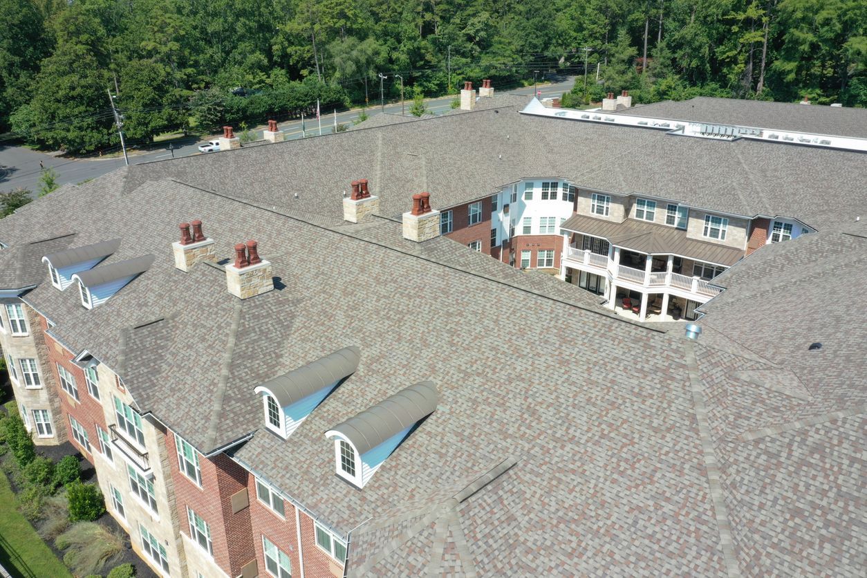 Aerial view of a multi-story building with a shingled roof, chimneys, and dormers; green trees in the background.