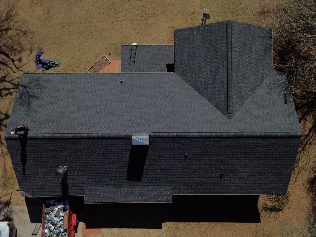 Aerial view of a dark gray shingled roof with workers, equipment, and a chimney on a sunny day.