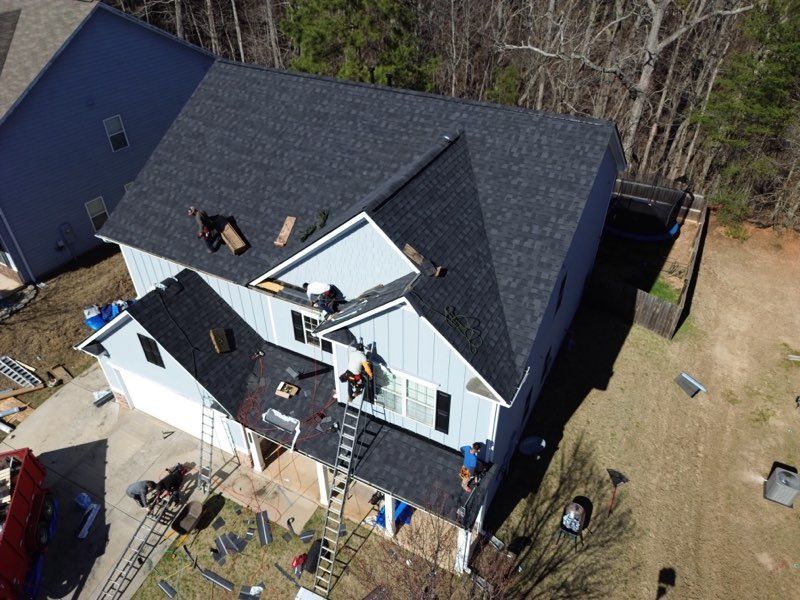 Overhead shot of house roof being repaired; workers on ladders with new shingles; light blue siding, black roof.
