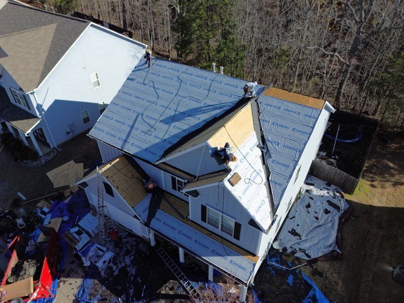 Aerial view of a house undergoing roofing repairs; workers on the roof, partially covered with blue underlayment and plywood.