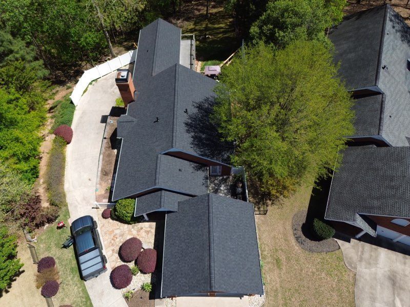 Overhead view of a house with a dark roof, surrounded by trees, a driveway, and a parked car.