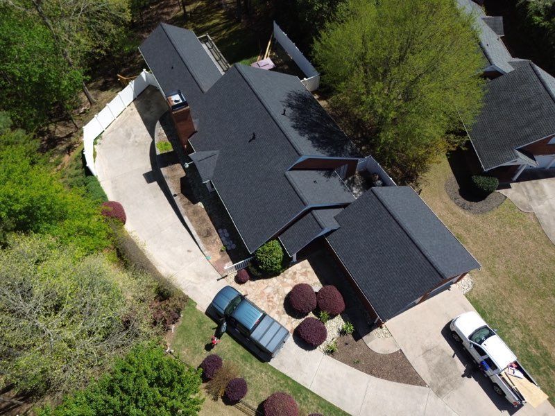Aerial view of a dark-roofed house with two vehicles in the driveway; surrounded by trees and landscaping.