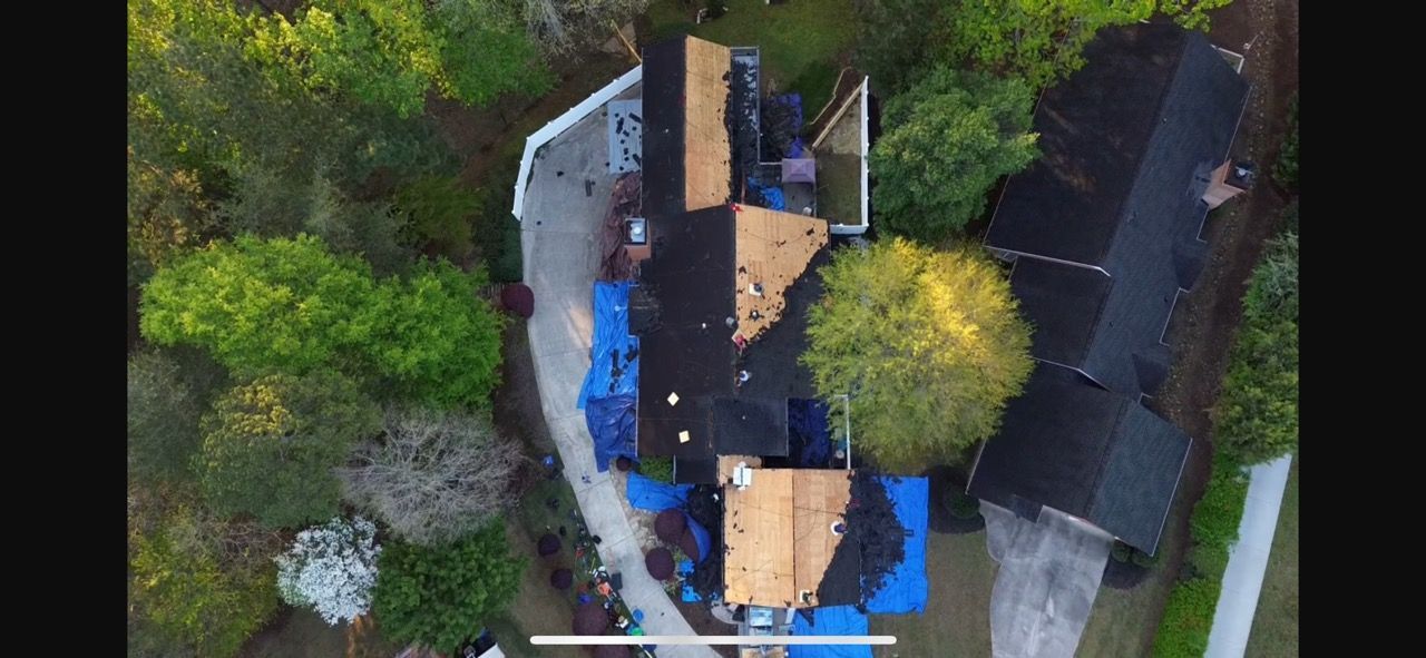 Aerial view of a damaged roof on a house. Blue tarps cover parts of the roof, surrounded by trees.