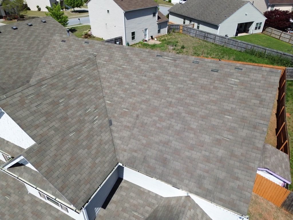 Overhead view of a house with gray shingles on the roof, surrounded by grassy yards and other houses.