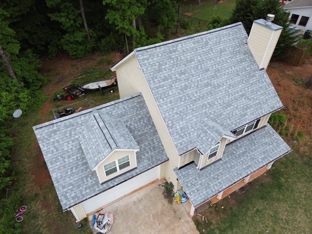 Aerial view of a beige house with a gray shingle roof, chimney, and a concrete driveway.