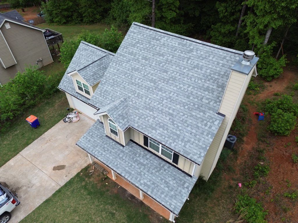Aerial view of a light yellow house with a gray roof and chimney, located in a wooded area.