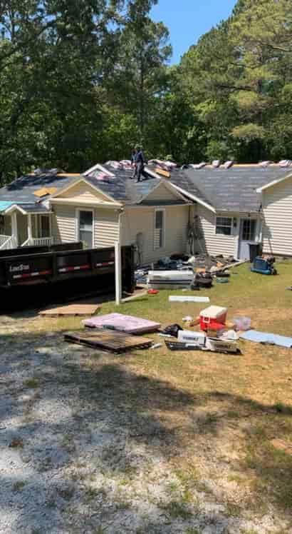 Roofers working on a house with a dumpster and materials on the ground in a sunny setting.