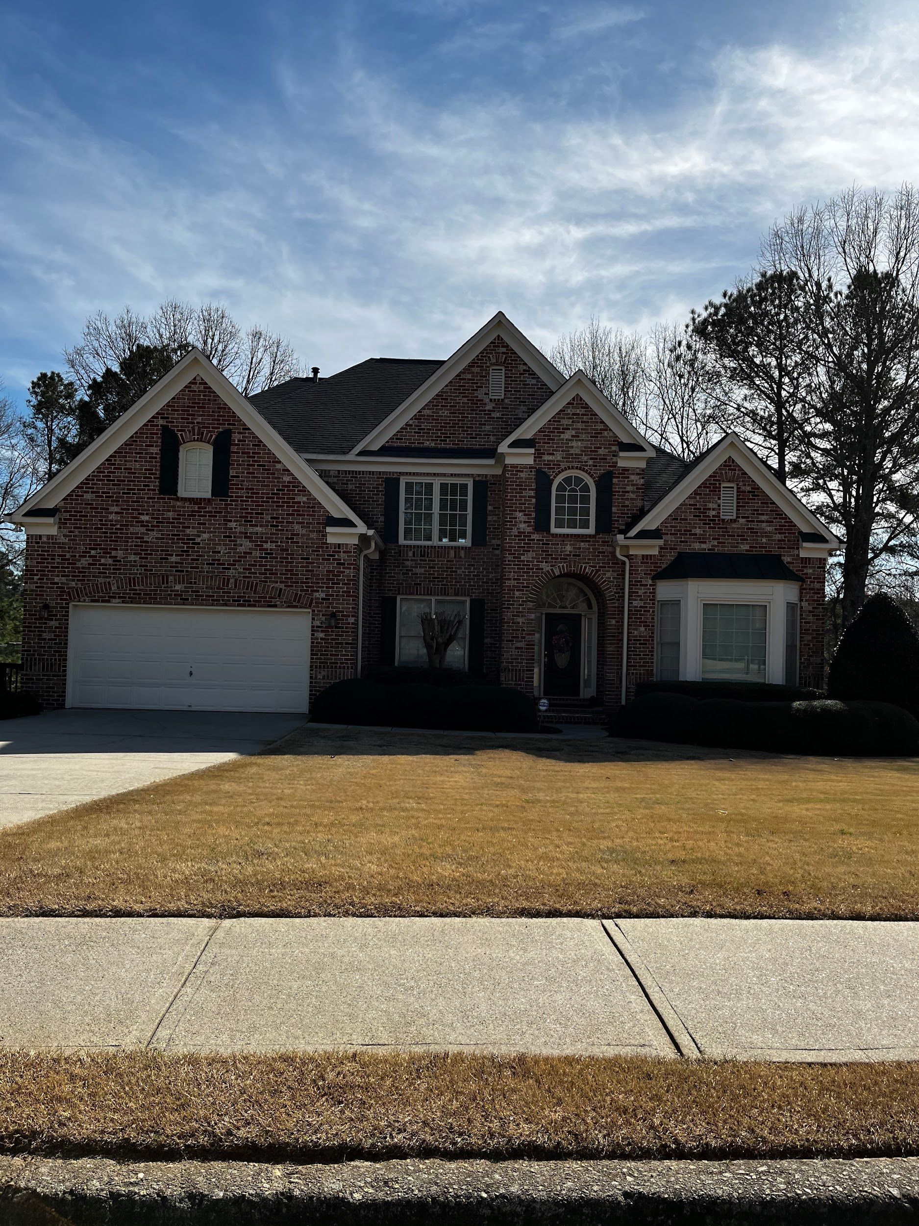 Two-story brick house with a white garage door and dry brown grass under a blue sky.