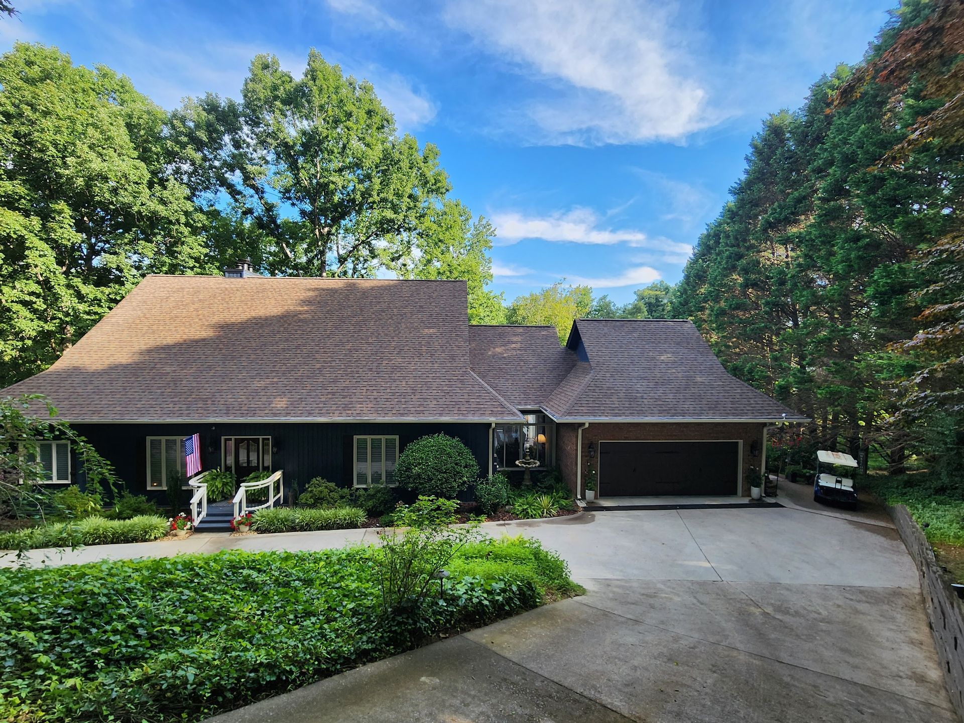 Dark house with brown roof and attached garage, surrounded by lush green trees under a blue sky.