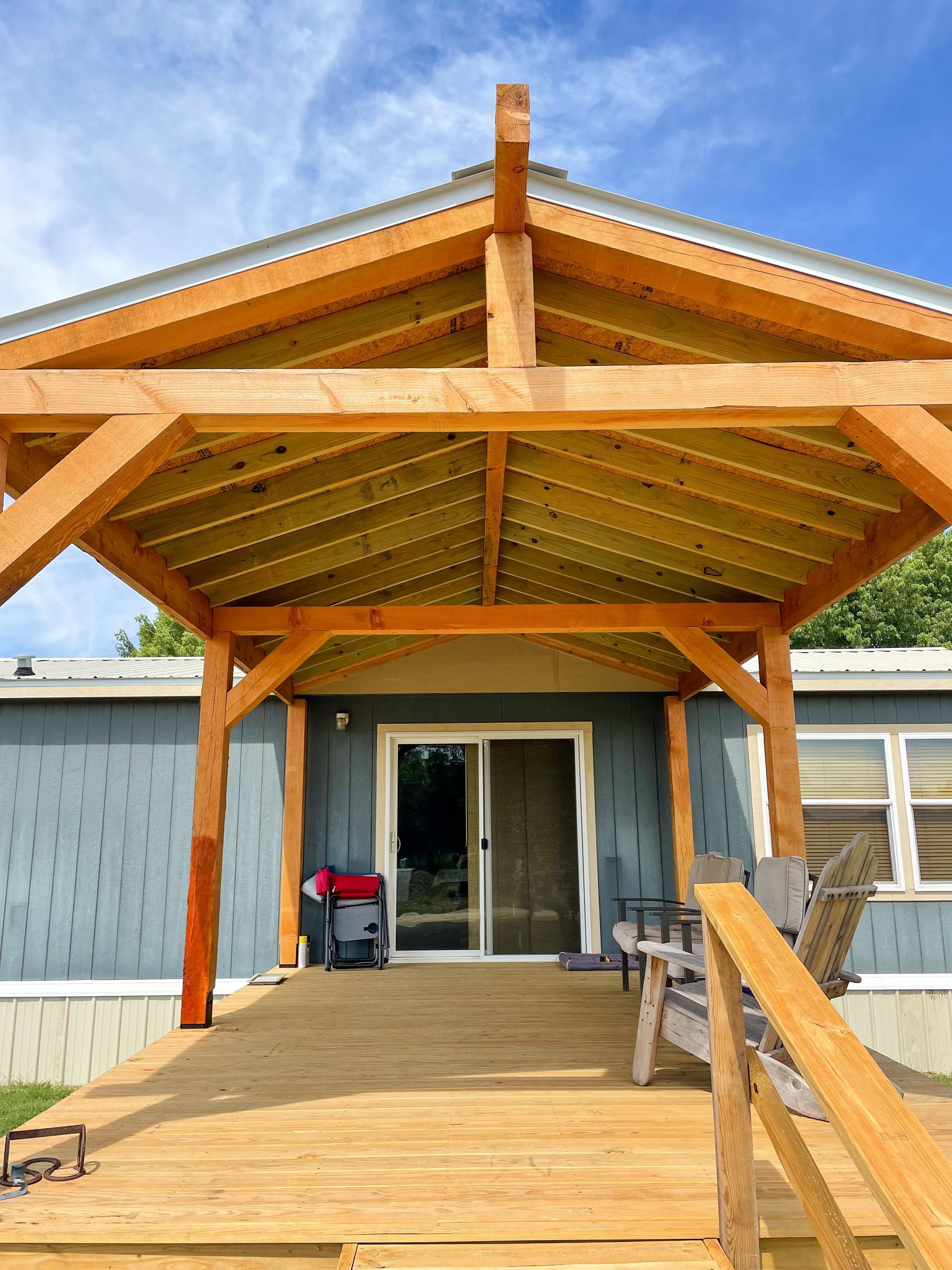 Wooden deck with a timber-framed porch roof over a sliding glass door; blue siding, clear sky.